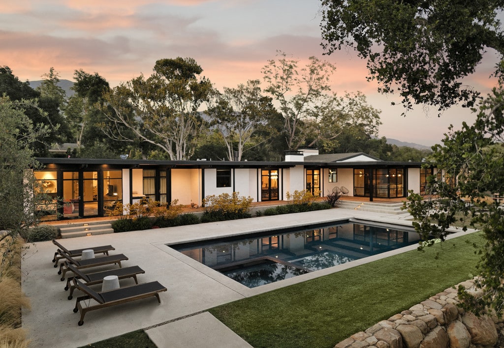 A sleek mid-century home designed by Lutah Maria Riggs, featuring clean lines and a minimalist aesthetic. The house has large windows, a flat roof, and an open floor plan. In the backyard, there is a sparkling blue pool surrounded by a well-manicured landscape, creating a perfect outdoor oasis.