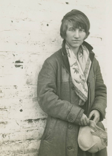 Black and white photo of Lutah Maria Riggs, an influential architect, standing against a wall. She is dressed in a coat, with a fashionable scarf draped on her shoulders, and holds a construction hat. This image highlights Lutah Maria Riggs' professionalism and hands-on approach to architectural projects.