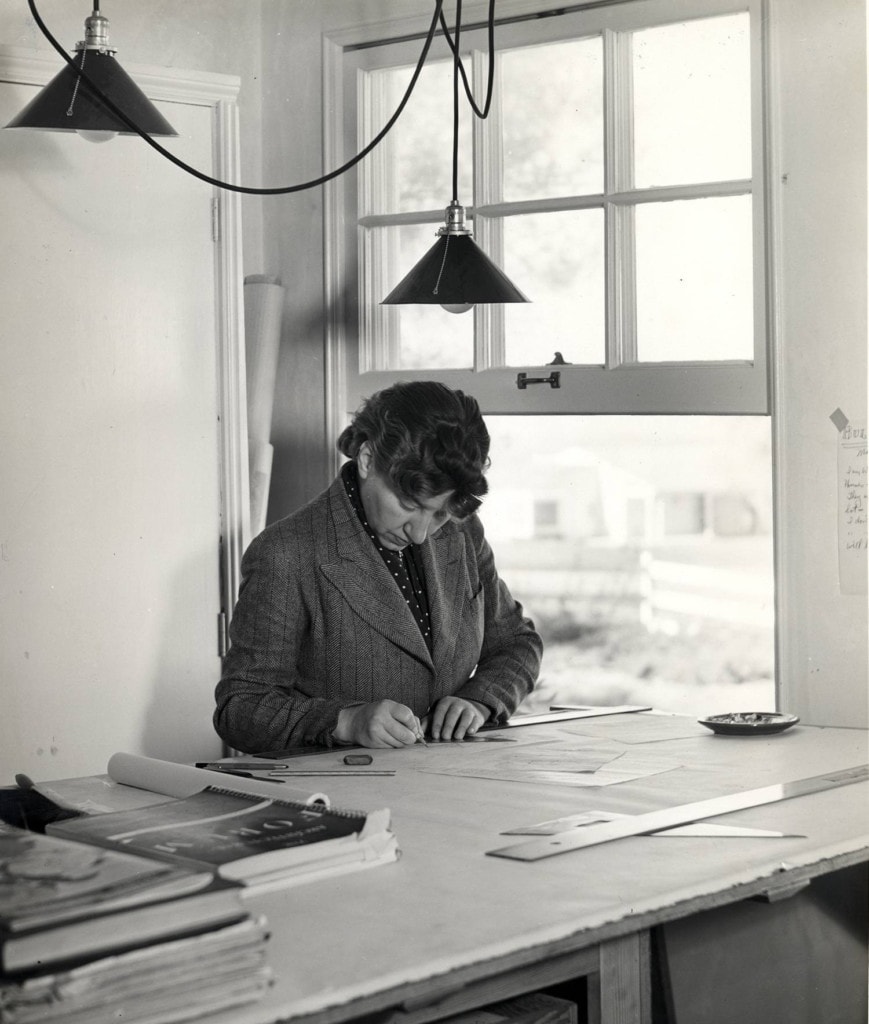 A black and white photograph of Lutah Maria Riggs at her desk, engrossed in her work. She is seated in a chair, leaning forward with focused concentration, examining architectural plans spread out on the desk in front of her.